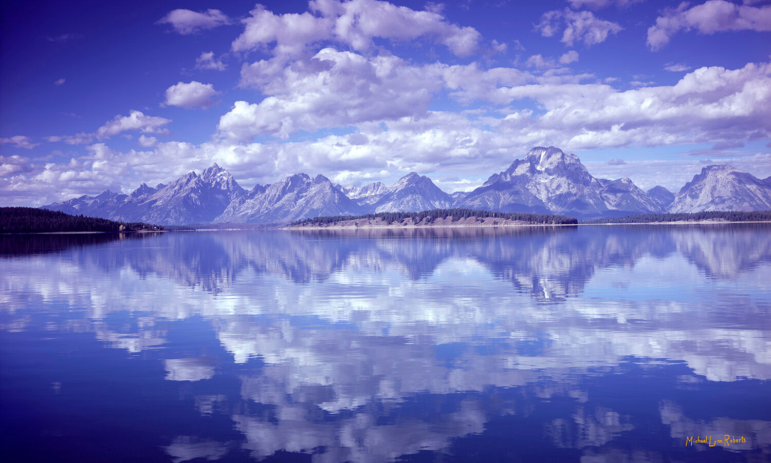 The Grand Teton Mountain range reflected in Jackson Lake along with a sky full of puffy white clouds.